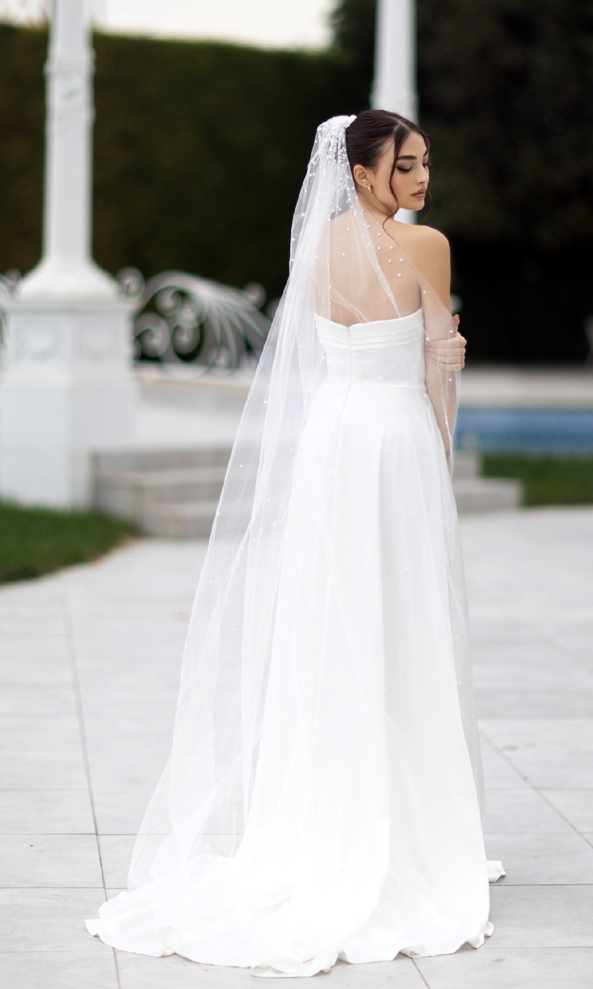 A woman wears the Off-White Strapless Bridal Gown: Tina Holly TW105 and a long veil outdoors, standing with her back to the camera and her head turned to the side.